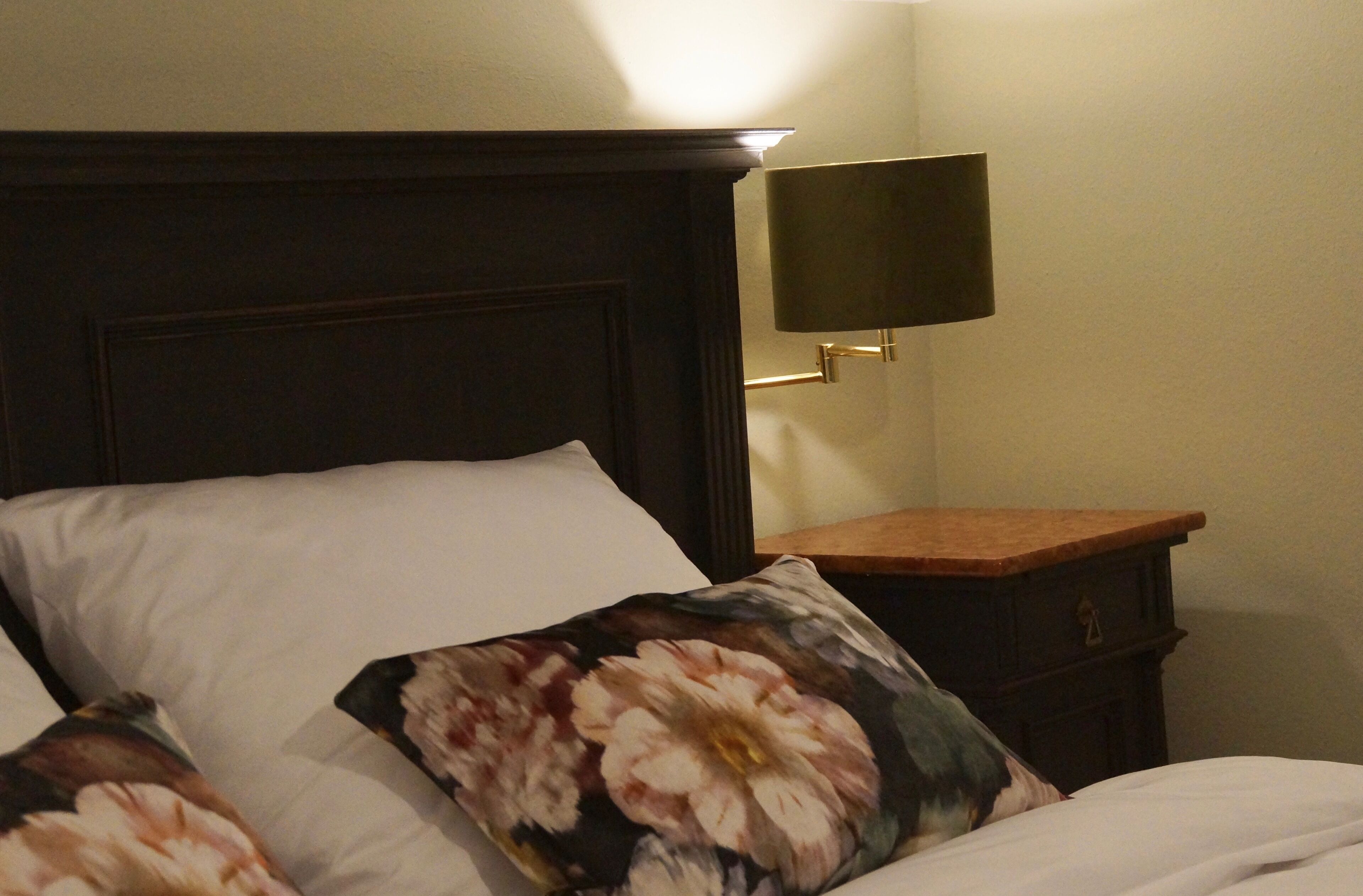 Habitación de hotel con cama, almohadas blancas, almohada estampada con motivos florales, cabecero oscuro, mesita de noche con lámpara y mesita auxiliar de madera.