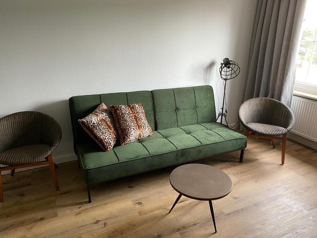 Un sofà verd amb coixins estampats de llebrera, dues cadires de vimetla i una taula rodona petita en una habitació amb terres de fusta i una làmpada de peu.