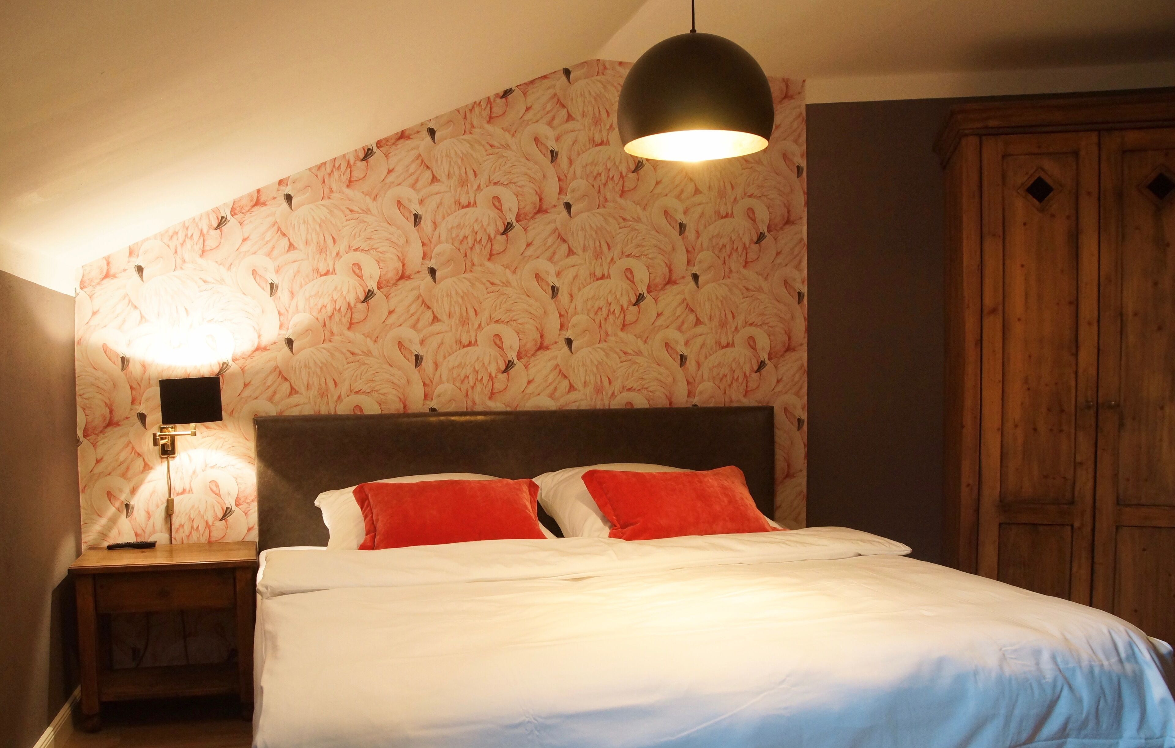 Un dormitorio con cama, mesita de noche, lámpara, armario de madera y pared con estampado de flamenco.