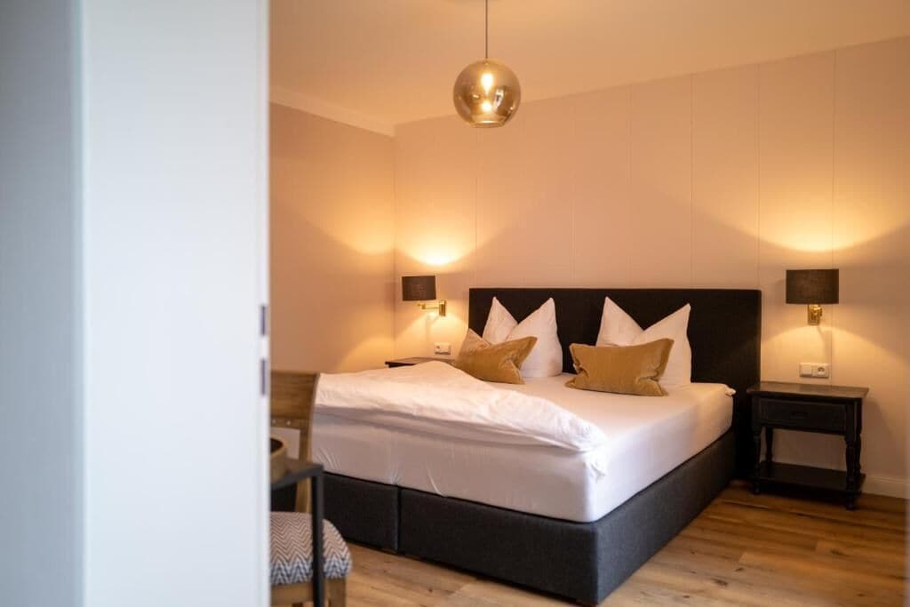 A neatly made bed with white linens and two pillows, a round pendant light above, and two bedside tables with lamps.