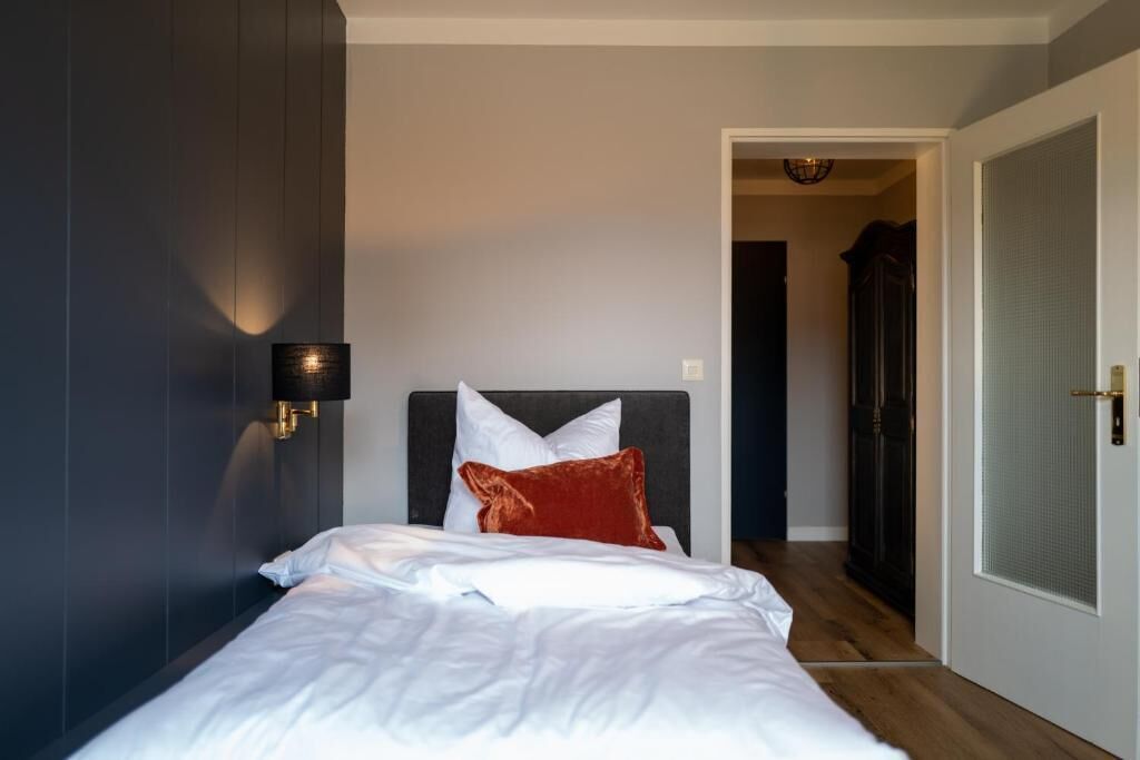 A bedroom with a bed, a nightstand, a wall-mounted lamp, and a door leading to another room.