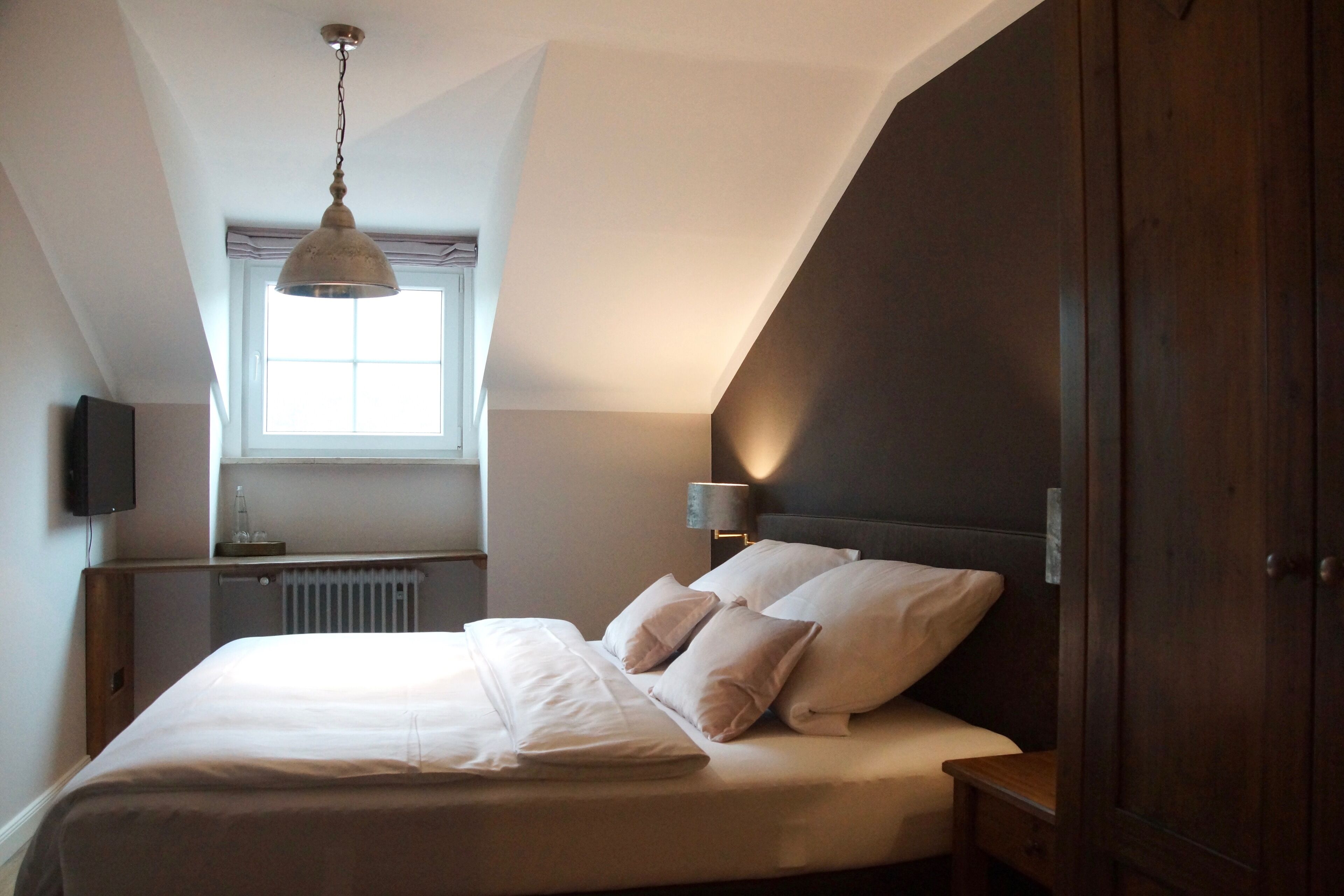 Een slaapkamer met een bed, nachtkastje, lamp, raam en een donkerhouten garderobe.