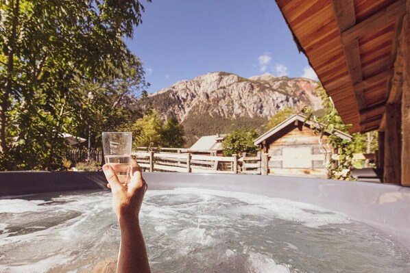 Un jacuzzi con montañas al fondo, construcciones de madera y cielo despejado.