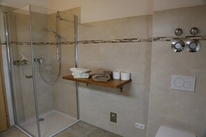 Une salle de bain moderne avec une douche à parois de verre, une étagère en bois où sont posées des serviettes et des toilettes dotées d’une poignée de chasse d’eau.