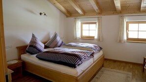 Uma cama de solteiro com roupa de cama listrada em um quarto com vigas de madeira no teto, duas janelas com cortinas e uma mesinha de cabeceira.