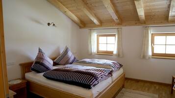 Ein Einzelbett mit gestreifter Bettwäsche in einem Zimmer mit Holzbalkendecke, zwei Fenstern mit Vorhängen und einem kleinen Nachttisch.