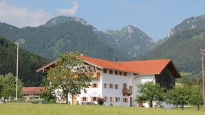 Um edifício de dois andares com telhado de telhas vermelhas, cercado por vegetação exuberante e montanhas ao fundo.
