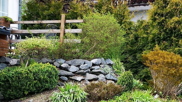 Ein angelegter Garten mit einer Steinsäule, verschiedenen Sträuchern und einem Holzzaun.