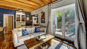 A cozy living room with a wooden ceiling, a white sofa with blue cushions, a wooden coffee table, and a large window with curtains.