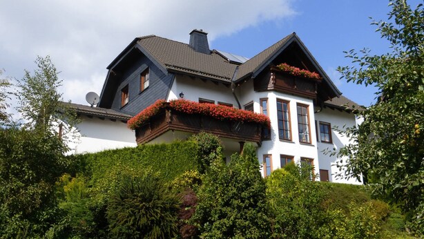 A large, two-story house with a steeply pitched roof, dormer windows, and a chimney. The house is surrounded by lush greenery and has flower boxes on the balcony. - Ferienwohnung Dunja (Winterberg)