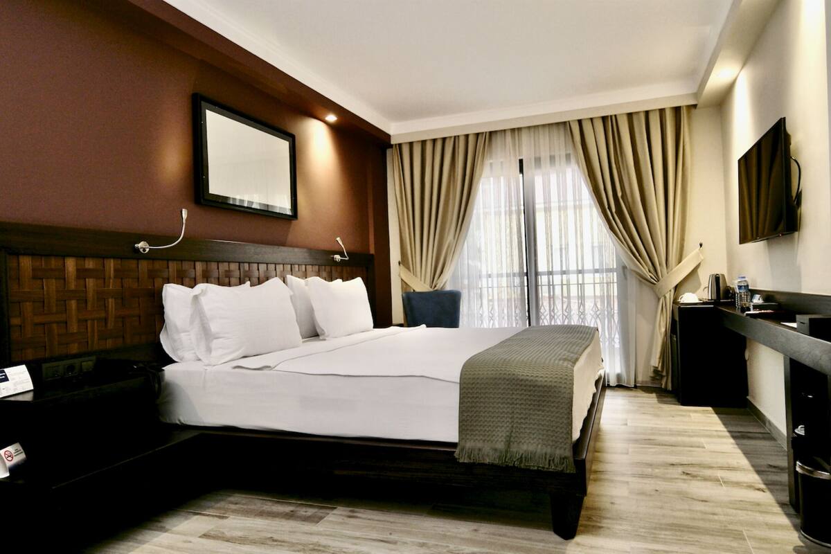 Habitació d'hotel amb un llit gran, un panell de capçalera de fusta, una taula de nit, un escriptori amb una cadira, un mirall i una finestra amb cortines.
