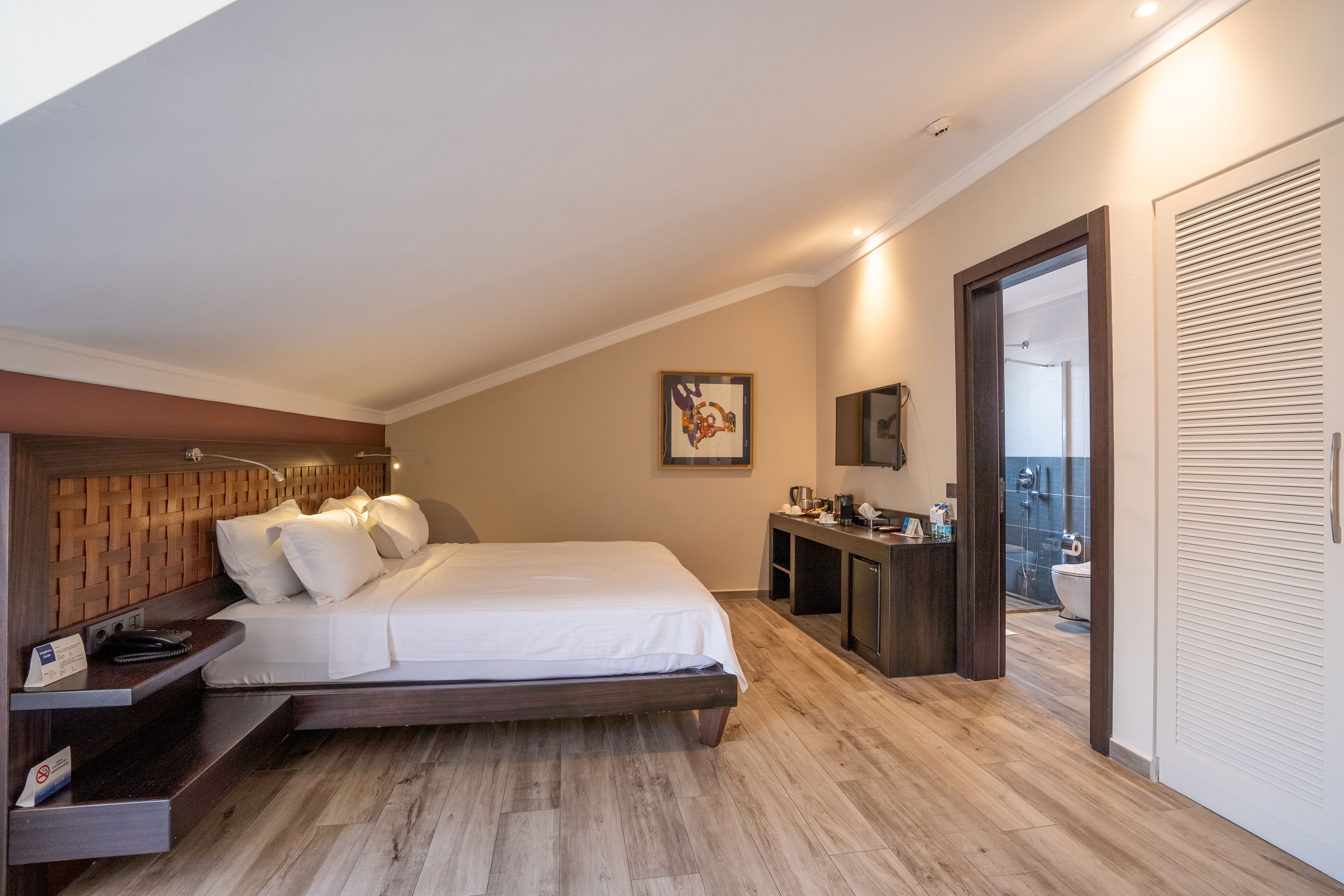 A modern hotel room with a wooden headboard, a large bed, a desk with a chair, a small bathroom, and a balcony with a view.