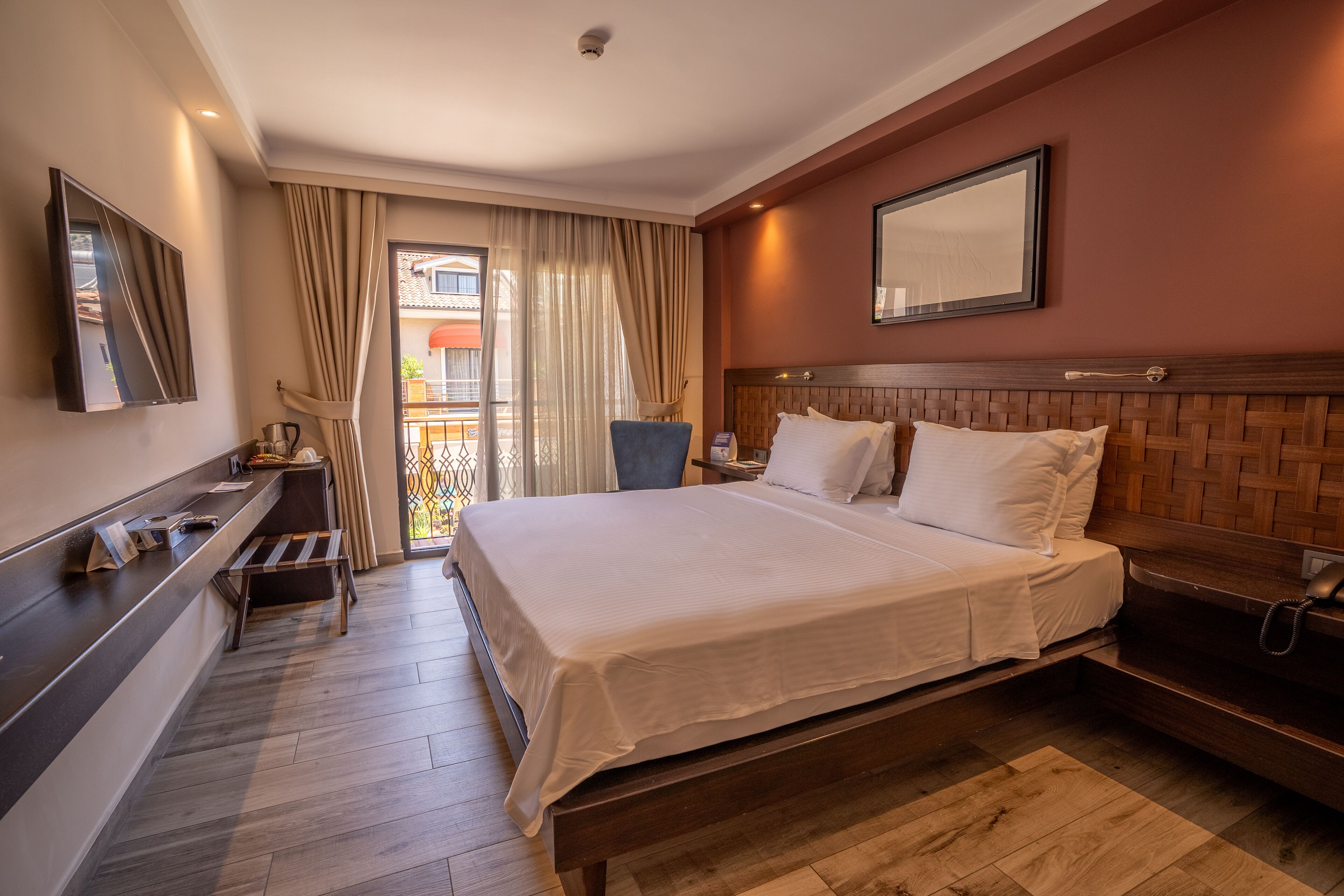 A hotel room with a large bed, a desk with a telephone, a chair, and a view of the outside balcony.