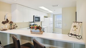 A modern kitchen with white cabinets, a microwave, and a refrigerator. There is a decorative basket with fruits and a small bird figurine on the counter. - Seacrest 504 is a Gulf Front 2 BR on Okaloosa Island by RedAwning (Fort Walton Beach)