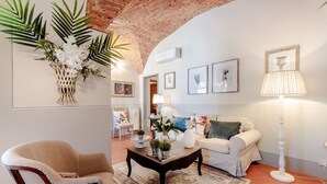 A living room with a white sofa, a wooden coffee table, and a beige armchair. There are framed pictures on the wall and a floor lamp.