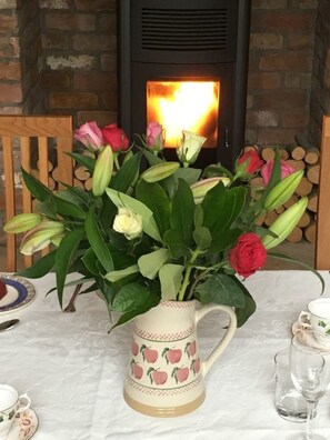 Una mesa con un arreglo floral en una jofaina, una chimenea y una pila de leña.