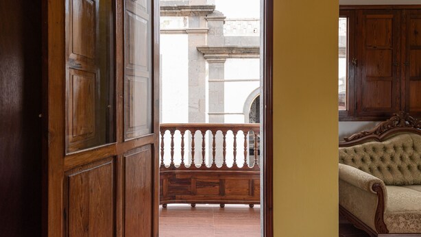 A wooden door leading to a balcony with a stone balustrade, a cushioned armchair, and a view of a stone building. - HomeForGuest NEW Duplex with Rooftop next to Arucas Cathedral (Arucas)