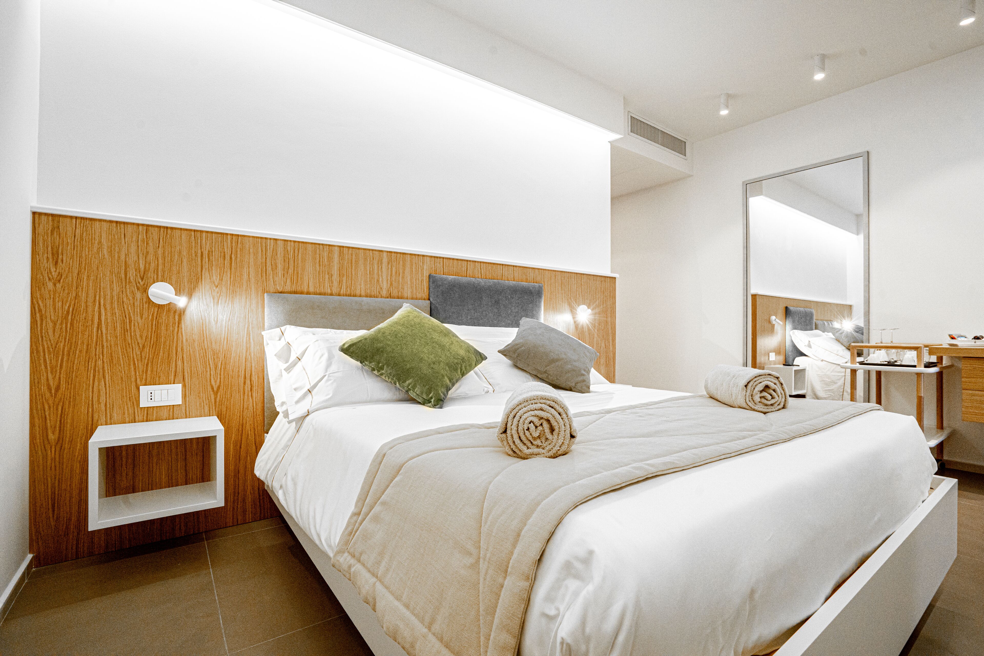 A modern hotel room with a wooden headboard, a bedside table, a mirror, and a neatly made bed with a rolled towel.