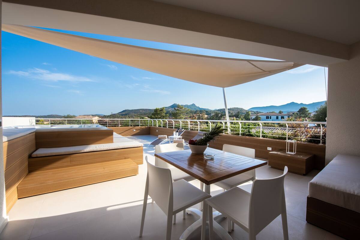 A balcony with a wooden table, white chairs, and a cushioned seating area.