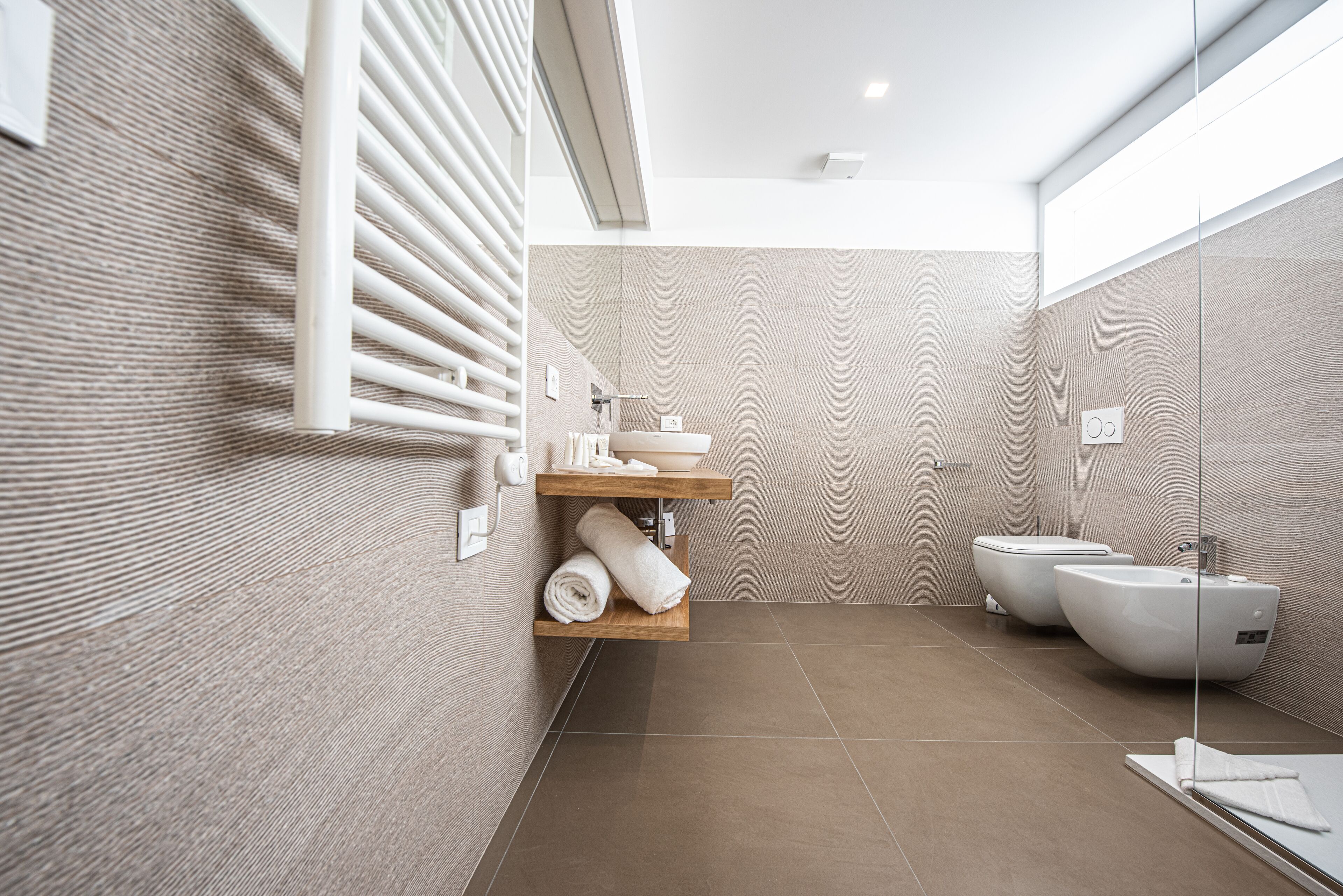 A modern bathroom with a glass shower enclosure, a wooden shelf with towels, and two white bidets.
