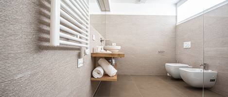 A modern bathroom with a glass shower enclosure, a wooden shelf with towels, and two white bidets.