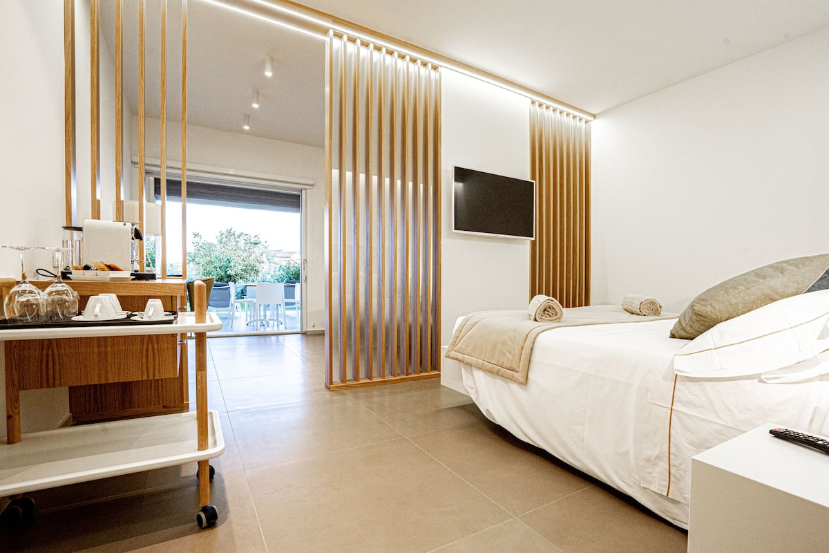A modern hotel room with a bed, a TV, a desk with a lamp, and a sliding door leading to a balcony.