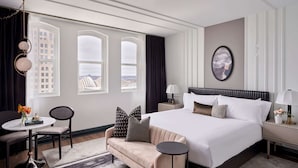 A hotel room with a large bed, a sofa, a small table, and a chair. There are two windows with curtains, a wall-mounted lamp, and a framed picture on the wall.