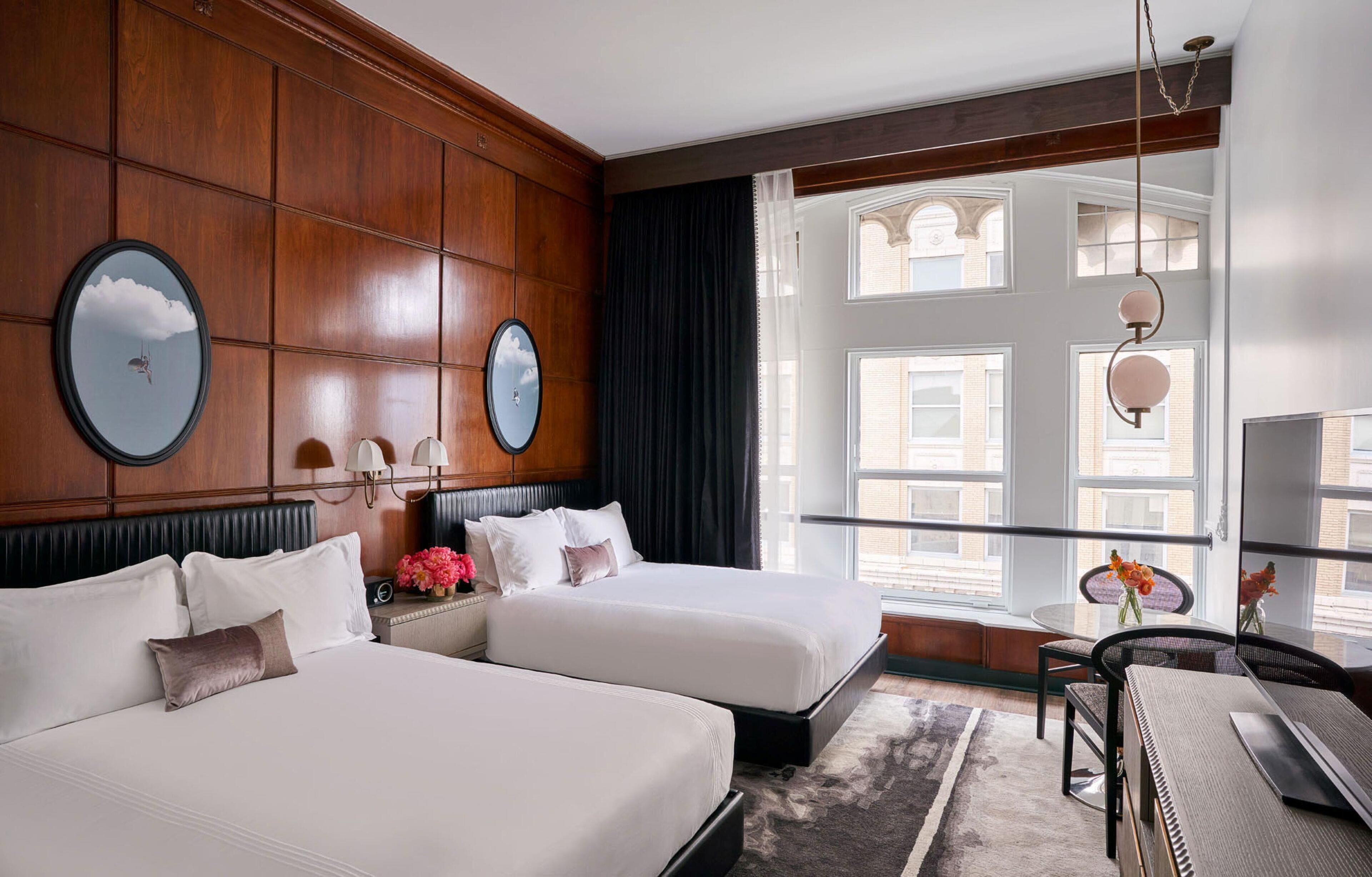 A hotel room with two beds, a desk, a chair, and a large window with a view of buildings.