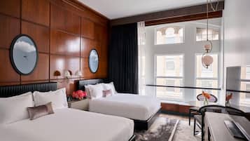 A hotel room with two beds, a desk, a chair, and a large window with a view of buildings.