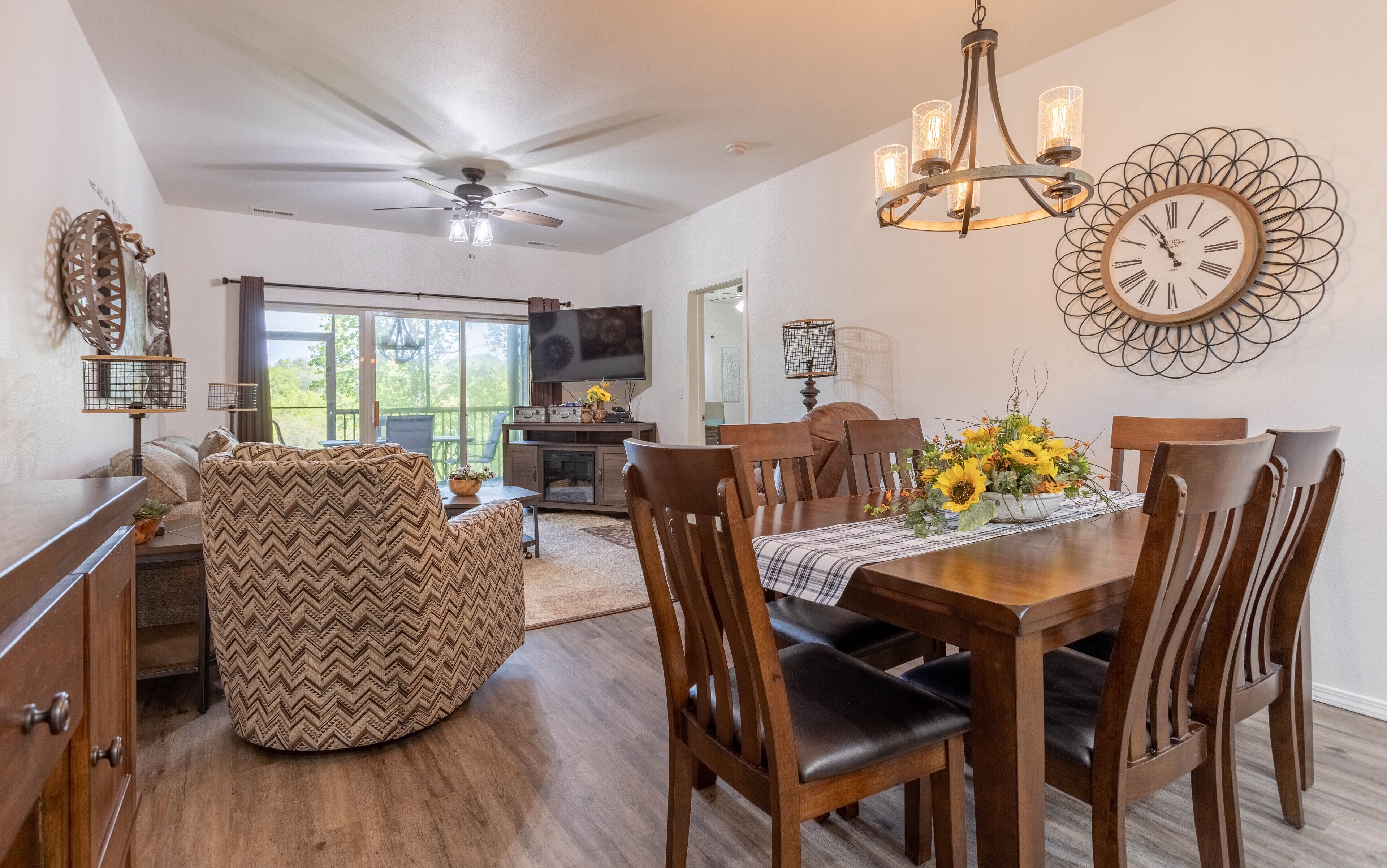 A dining area with a wooden table, chairs, and a chandelier. There is a television in the background and a ceiling fan.