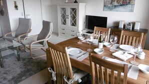 Comedor con una mesa de madera dispuesta para cuatro personas, rodeada de sillas de madera. Se observa un aparador con puertas de cristal, un televisor y un cuadro en la pared.