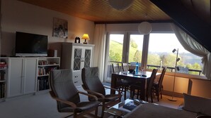 Una sala de estar acogedora con televisor, estanterías, un comedor con sillas y mesa, y un amplio ventanal con vistas panorámicas.