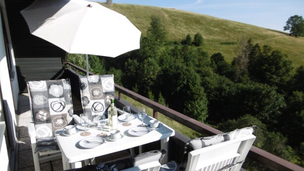 Un área de comedor al aire libre con mesa y sillas, una sombrilla y vistas a una ladera verde.