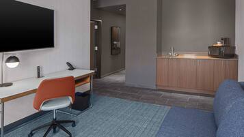A modern hotel room with a flat-screen TV, a desk with a chair, a kitchenette, and a blue sofa.