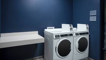 A laundry area with two front-loading washing machines, a white shelf, and two signs indicating laundry supplies and cycle times.