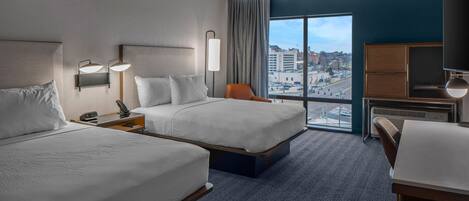 Ein Hotelzimmer mit zwei Betten, einem Schreibtisch, einem Stuhl und einem großen Fenster mit Blick auf die Stadt.