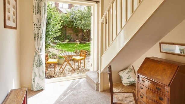 A room with a staircase, a wooden chest of drawers, a wicker chair, and a view of a garden through a glass door. - Host & Stay | Princes Road (Saltburn-by-the-Sea)