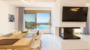Ein modernes Wohnzimmer mit großem Flachbildfernseher, einem Essbereich mit Holztisch und Stühlen sowie Blick auf einen Swimmingpool durch eine Glastür.