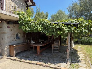 Eine Steinsitzbank unter einer mit Wein bepflanzten Pergola, eine rustikale Holzbank und eine Statue in einer Nische.