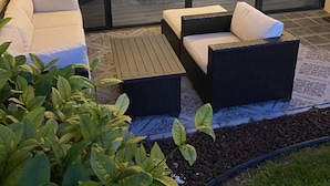 An outdoor seating area with cushioned sofas and a wicker table, surrounded by potted plants and a garden hose. - Newly Renovated Condo with SUP Paddle Boards and Golf Clubs Included (Kailua-Kona)