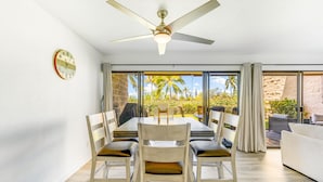 A dining area with a table and chairs, a ceiling fan, and a view of palm trees through large windows. - Newly Renovated Condo with SUP Paddle Boards and Golf Clubs Included (Kailua-Kona)