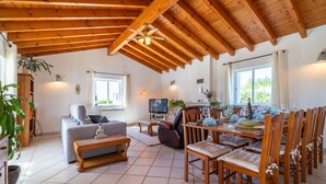 Een ruime woonkamer met een hoog houten plafond, een bank, een eettafel gedekt voor zes personen en een televisie.