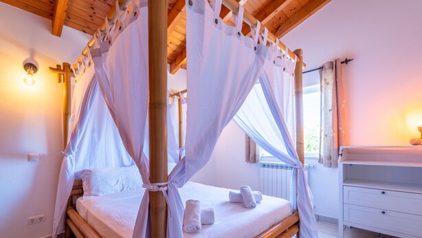 Een kamer met een bed, een houten plafond, een raam met gordijnen en een nachtkastje.