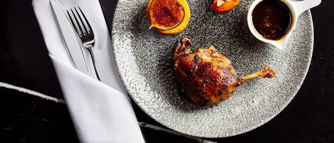 A plate with a roasted chicken leg, a halved orange, and a small bowl of sauce, accompanied by a fork and napkin on a dark surface.