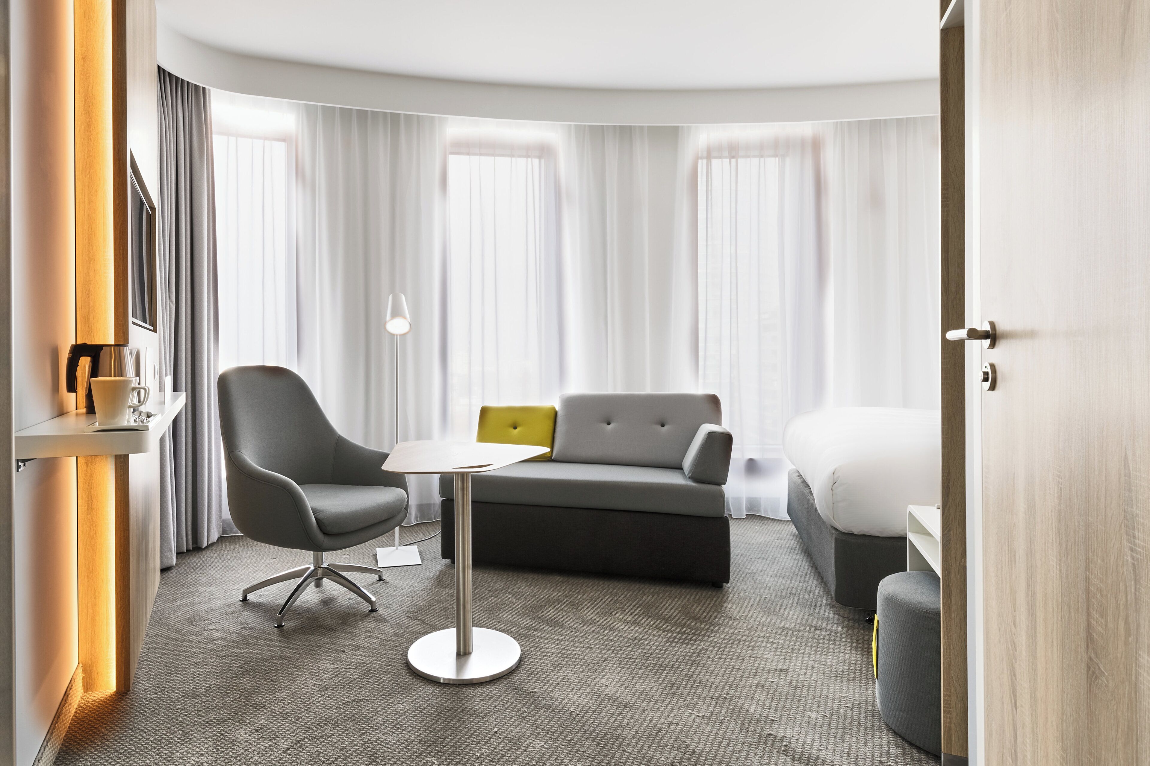 Ein modernes Hotelzimmer mit einem grauen Sofa, einem kleinen runden Tisch, einem einzelnen Sessel, einem Bett und einem Nachttisch mit Lampe.