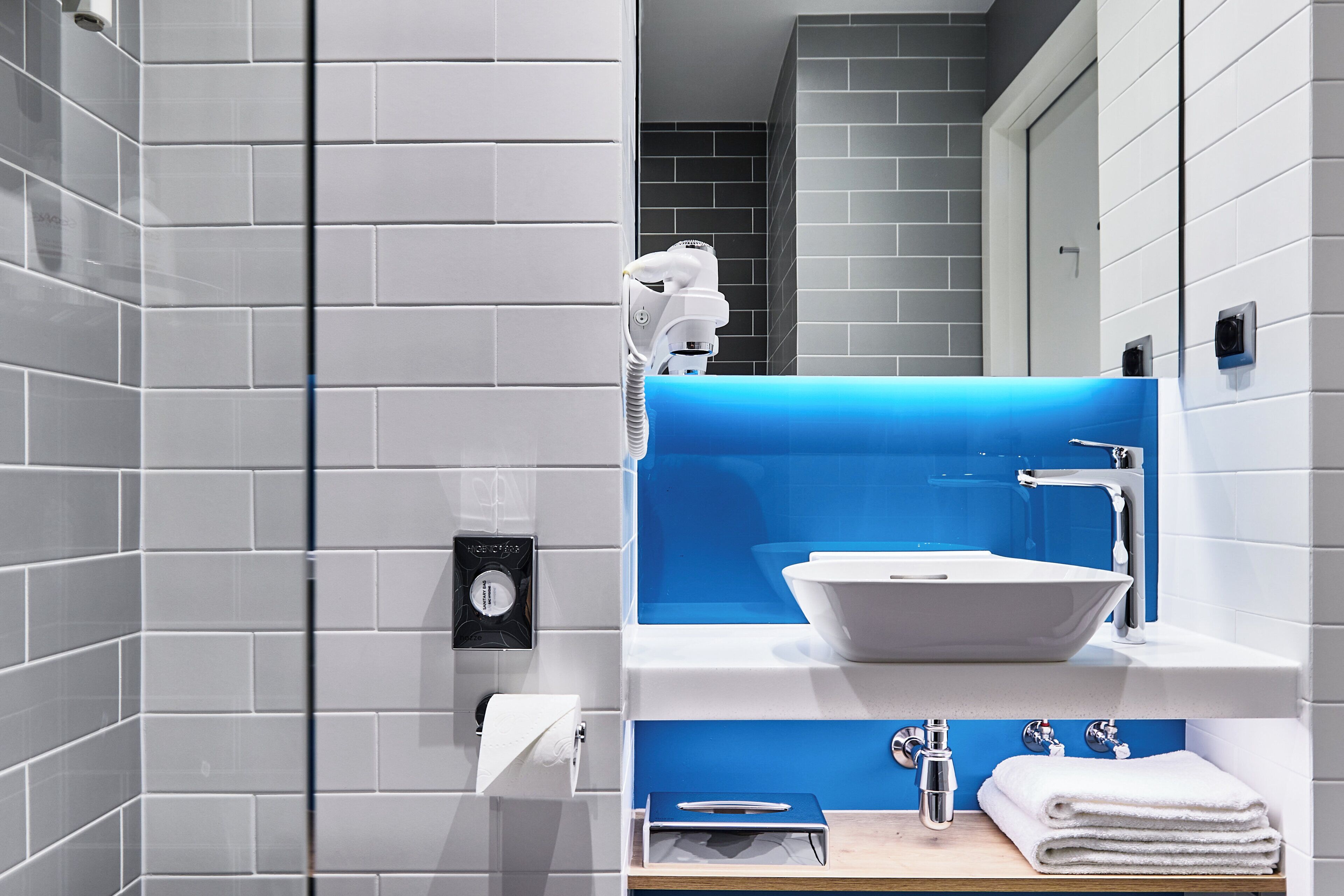 A modern bathroom with white tiles, a blue accent wall, a white sink, and a mirror.