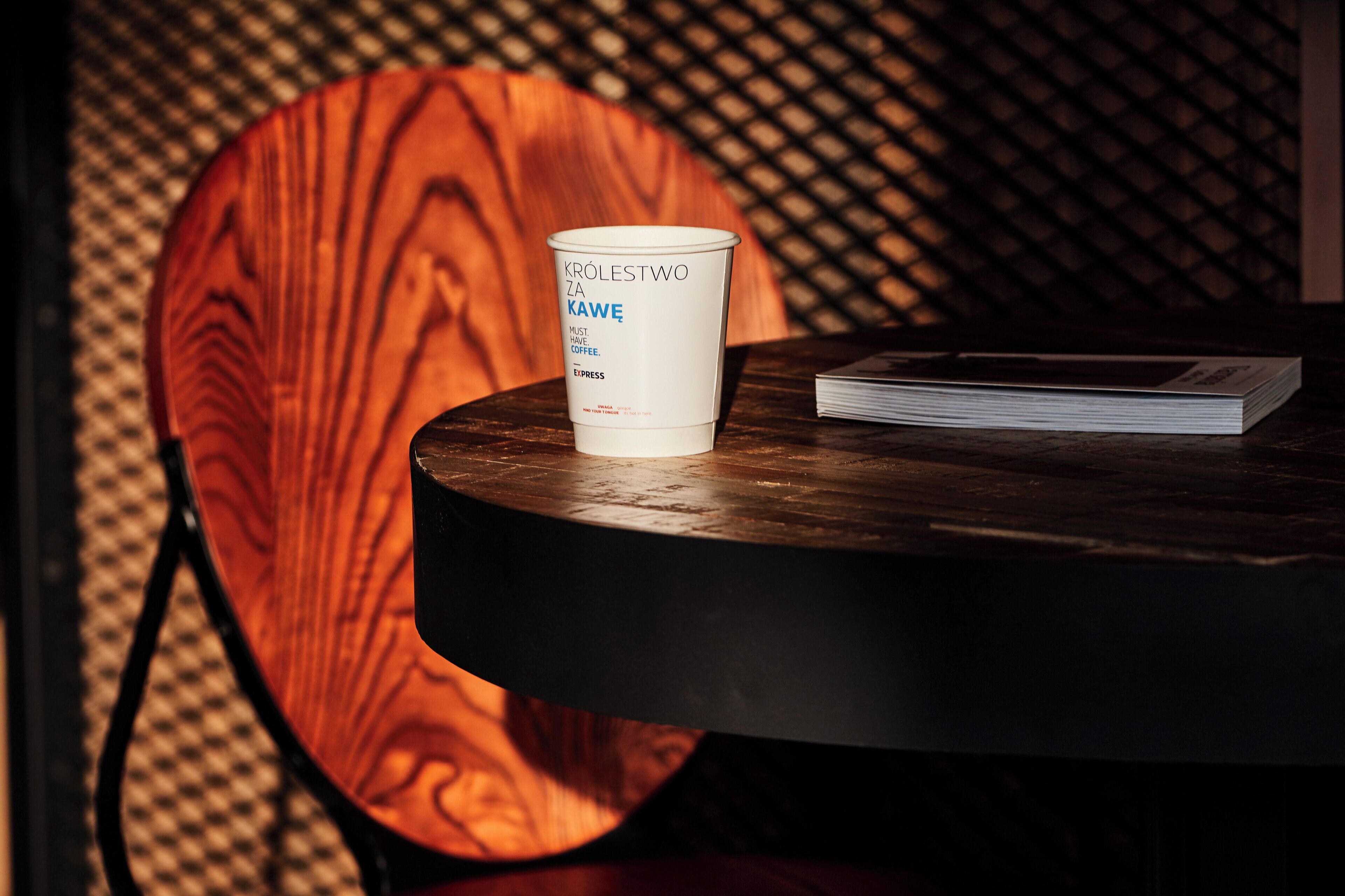 Une tasse à café blanche, posée sur une table ronde, avec du texte imprimé dessus, à côté d’un livre et d’une chaise en bois dont le dossier est en osier tressé.