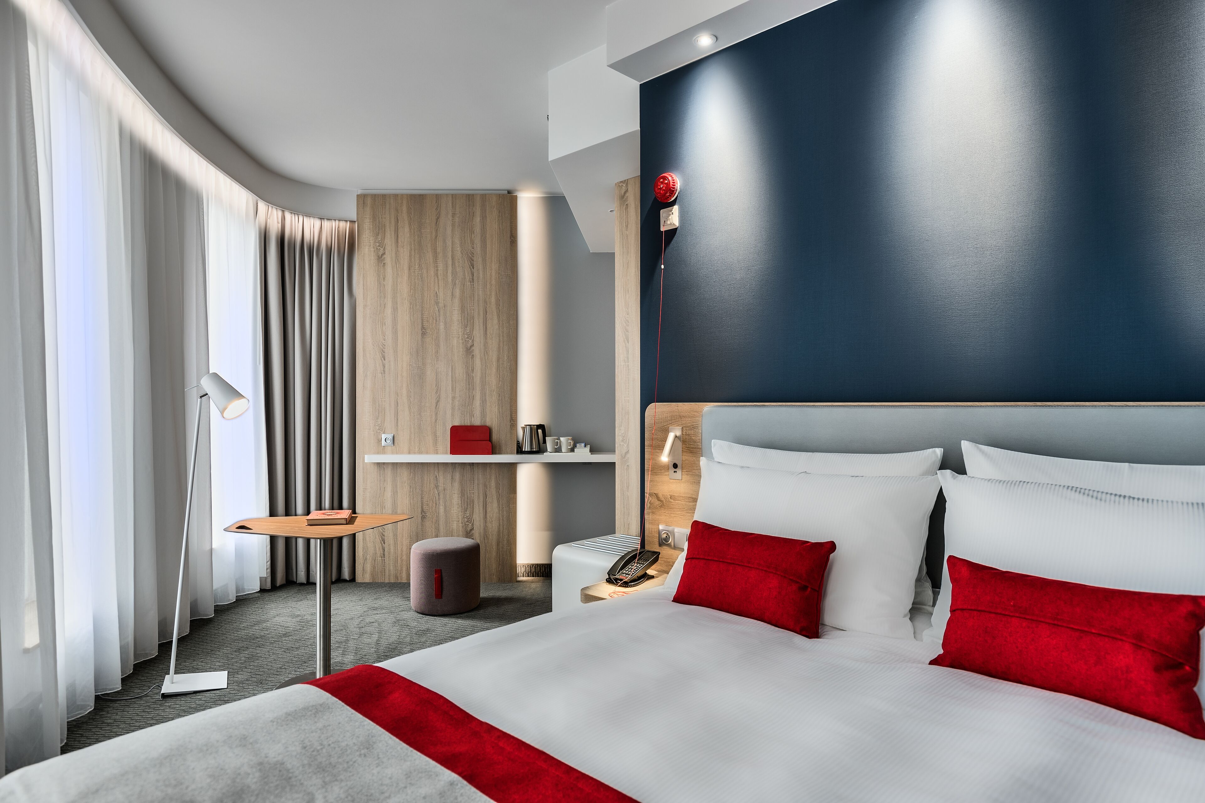 A modern hotel room with a large bed, a bedside table, a lamp, and a small stool.