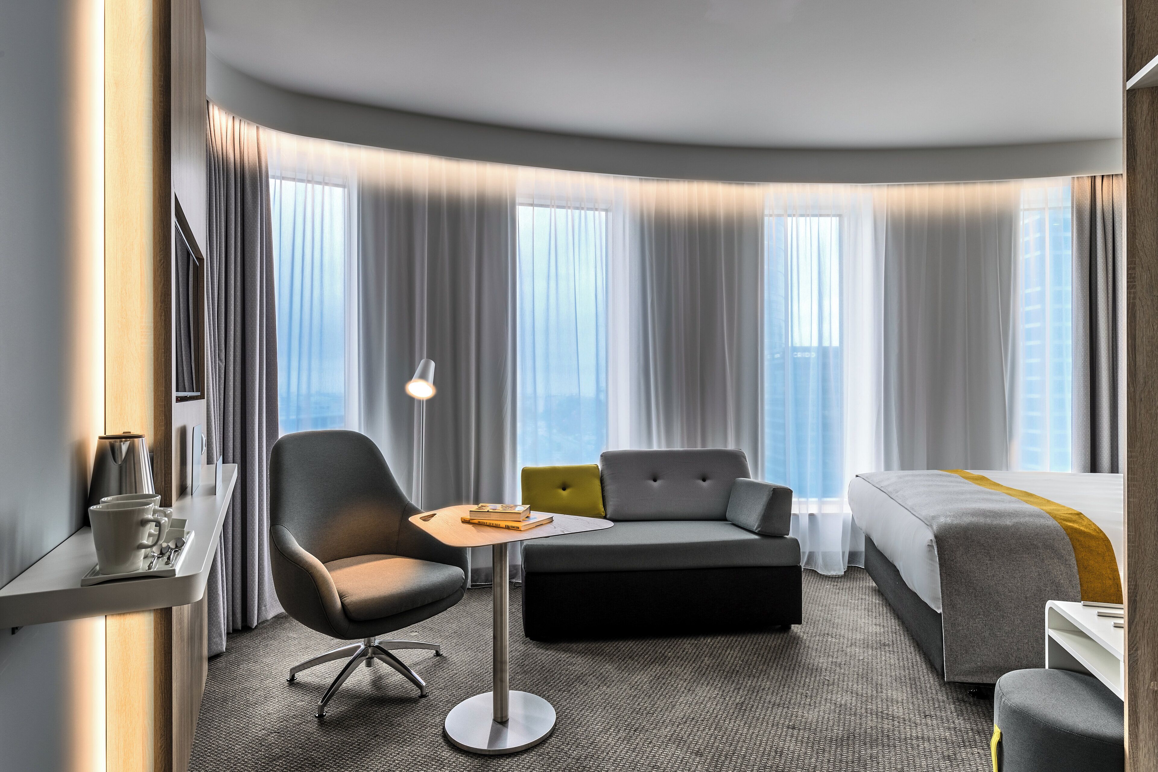 A modern hotel room with a curved bed, a small sofa, a desk with a lamp, and a view of the city through large windows.