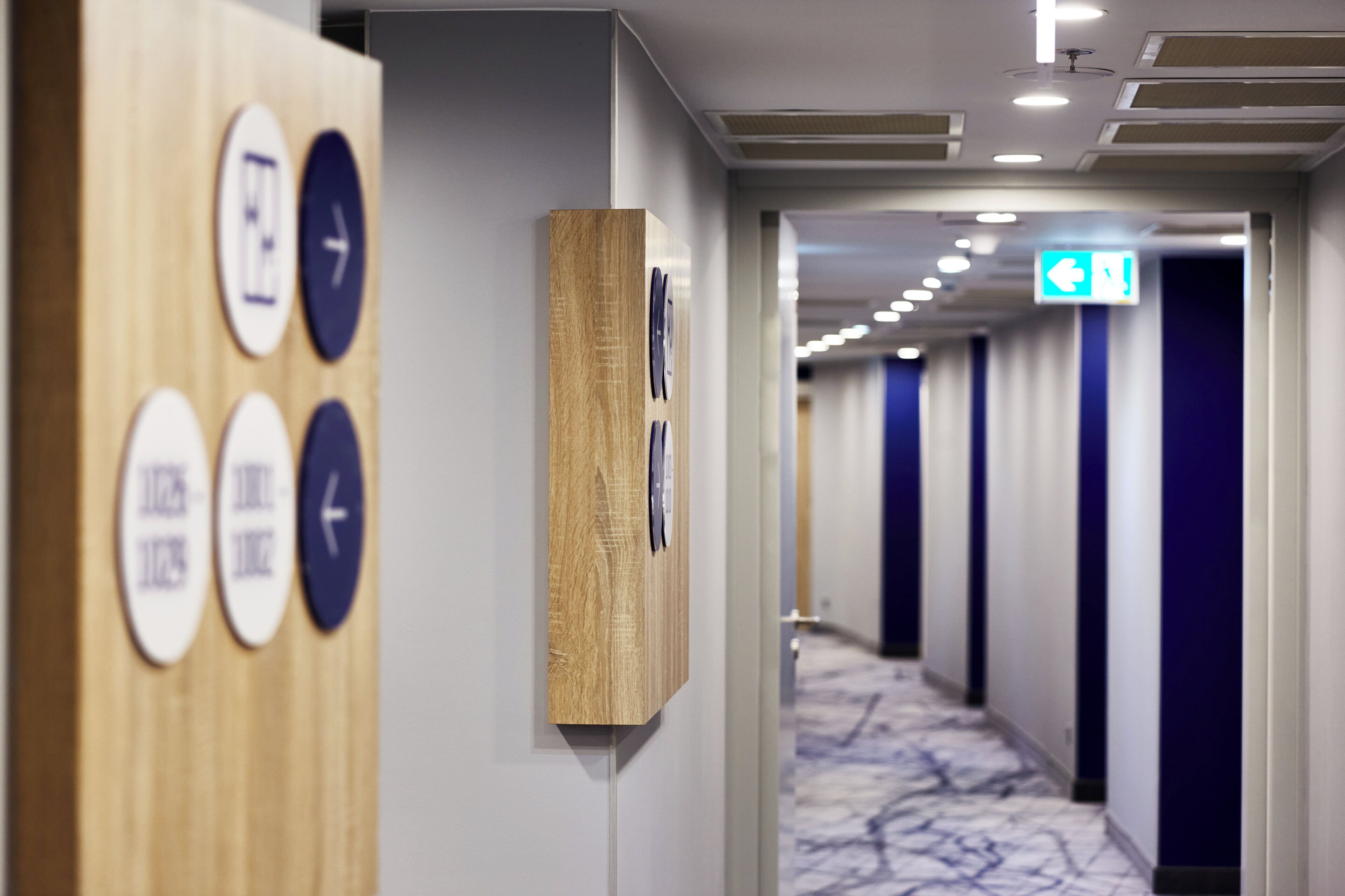 Un couloir d’hôtel avec des panneaux de signalisation au mur, un panneau décoratif en bois et un tapis à motifs.